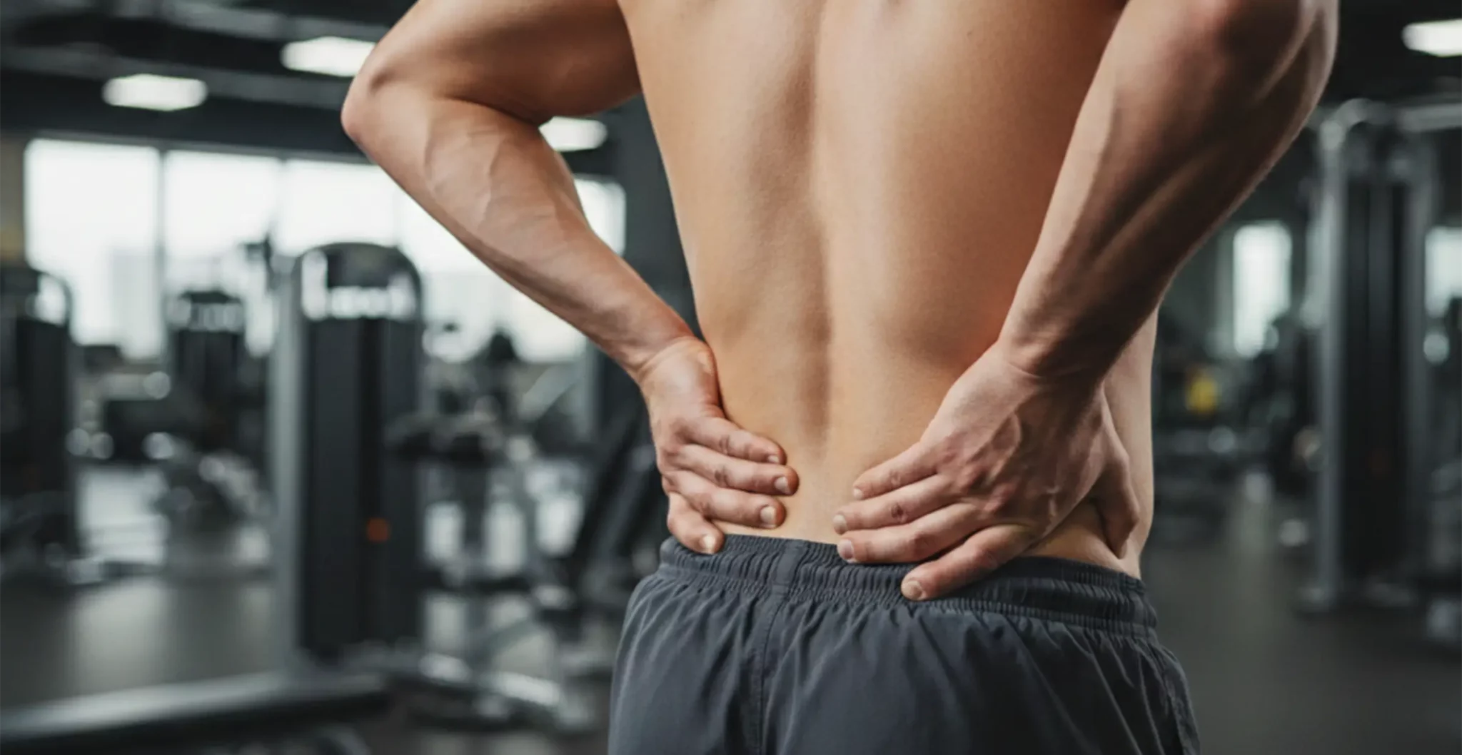 Homme sportif de dos, mains sur le bas du dos, illustrant une douleur lombaire liée à la musculation et à la hernie discale dans une salle de sport