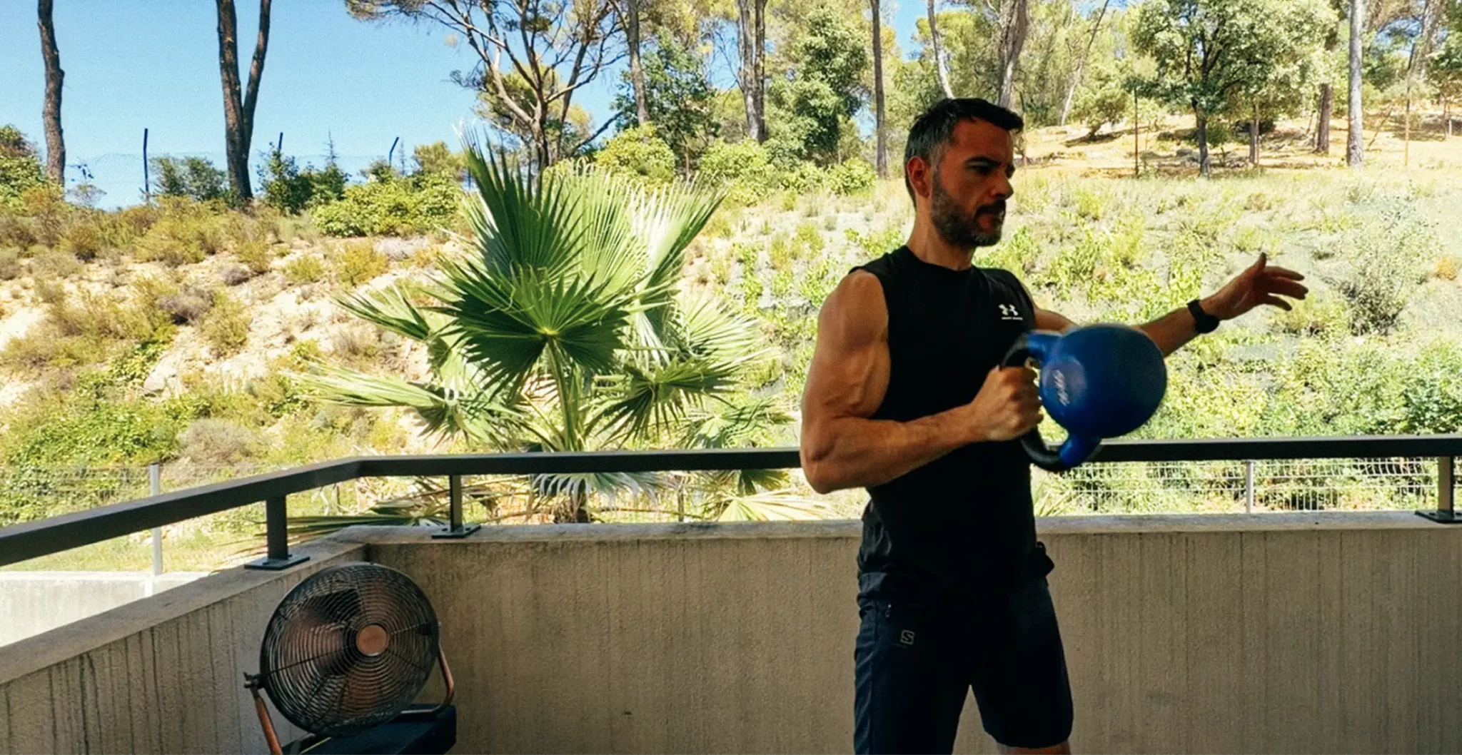Homme s’entraînant à la kettlebell en extérieur pendant une canicule, sur un balcon ombragé avec ventilateur
