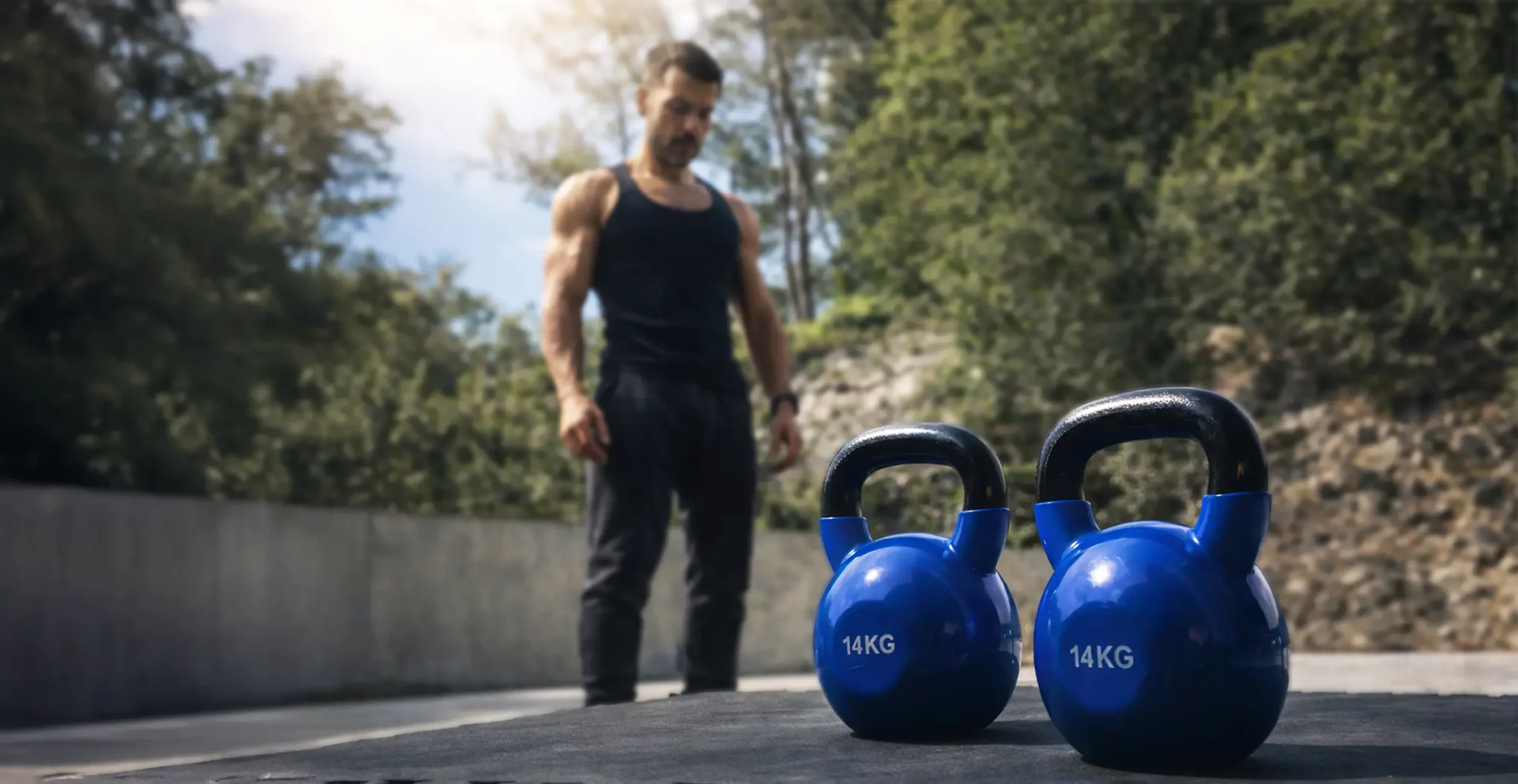 Homme musclé en débardeur noir devant deux kettlebells bleues de 14kg en extérieur - résultat entraînement kettlebell