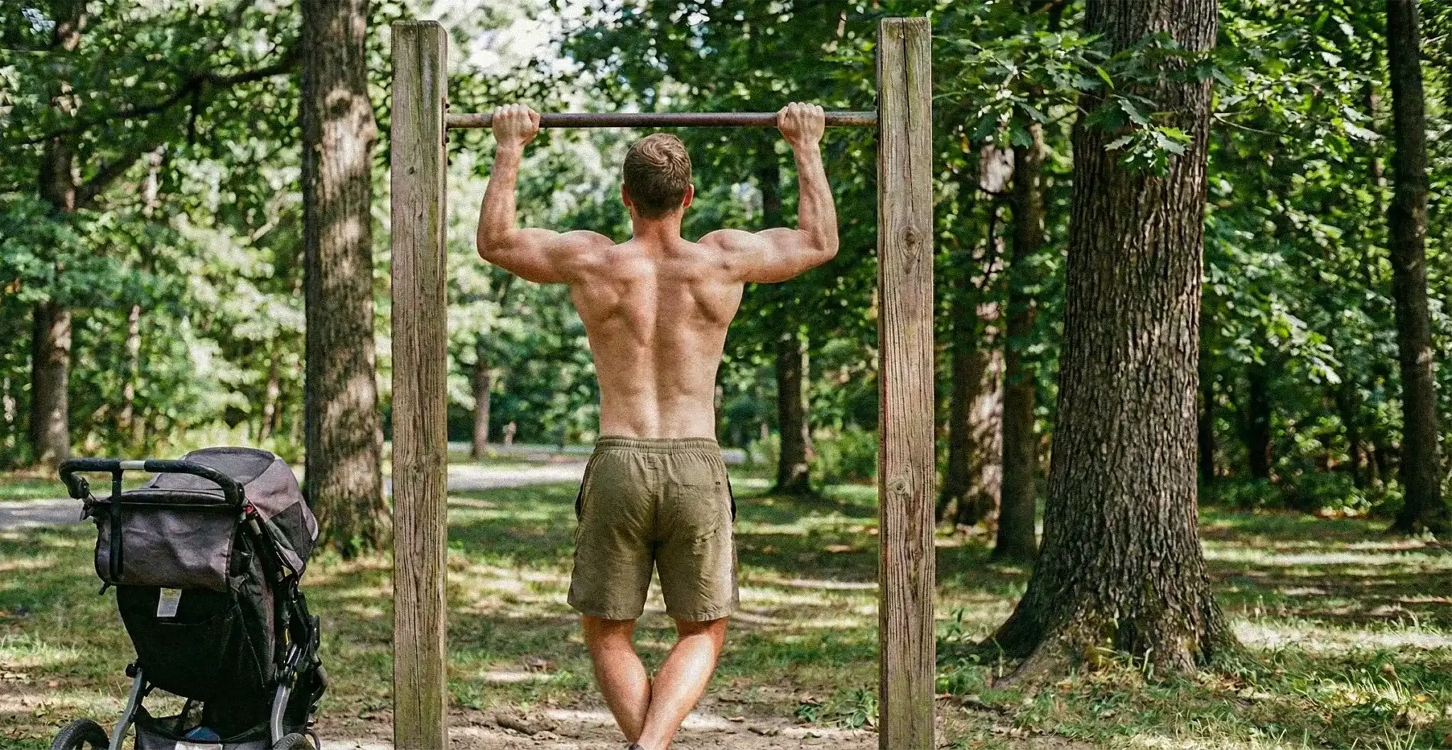 Jeune papa musclé effectuant des tractions sur une barre en extérieur avec une poussette de bébé garée à côté.