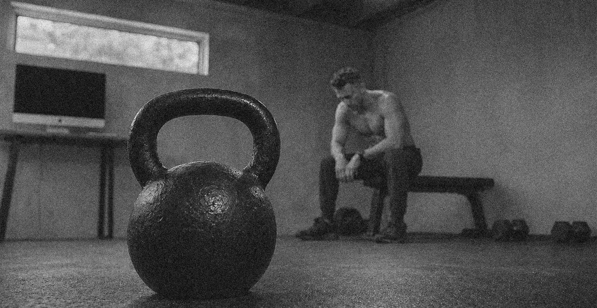 Homme assis seul sur un banc de musculation dans son home gym, kettlebell au premier plan, ambiance d'isolement liée au télétravail